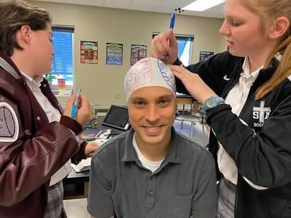A student draws the regions of the brain on a white cap on Paul’s head while he grins.