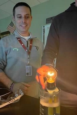 Paul Levy smiling in a classroom, holding a glowing light bulb over a beaker.